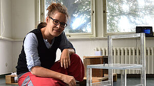 Ramona Seyfarth hockt vor einer weißen Platte. Darauf steht ein transparenter Kinderstuhl mit Lehne. Auf der Sitzfläche steht ein kleiner Spielzeugsoldat. Auf der Platte liegt eine bunte, geschmolzene Masse.