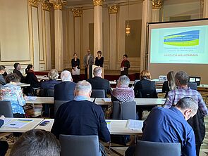 Ein Saal voller Menschen. Sie sitzen auf mehrere Reihen verteilt. Ein Beamer wirft eine Präsentation auf eine Leinwand. Am Rednerpult steht eine Frau und spricht.