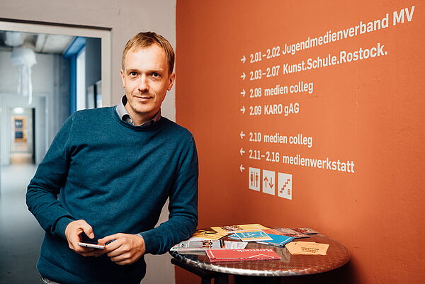 Hendrik Menzl lehnt an einem runden Stehtisch, auf dem Flyer liegen. Neben ihm steht eine orangefarbene Wand mit Wegweisern durch das Service-Center.