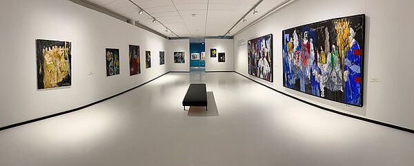 Ein Ausstellungsraum. An der Wand hängen Bilder. in der Mitte steht eine Sitzbank.