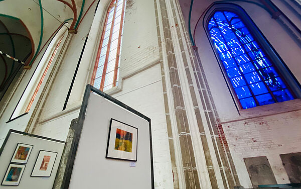 Links stehen Aufsteller, an denen Kunstwerke für die Versteigerung hängen. Rechts befindet sich ein hohes Kirchenfenster mit blau-weiß schattiertem Glas.  