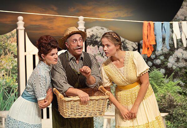 Ein Mann steht auf einer Bühne. Neben ihm stehen zwei Frauen. Hinter ihnen hängt Wäsche.