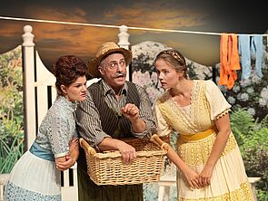 Ein Mann steht auf einer Bühne. Neben ihm stehen zwei Frauen. Hinter ihnen hängt Wäsche.