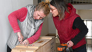 Zwei Frauen öffnen eine Holzkiste.