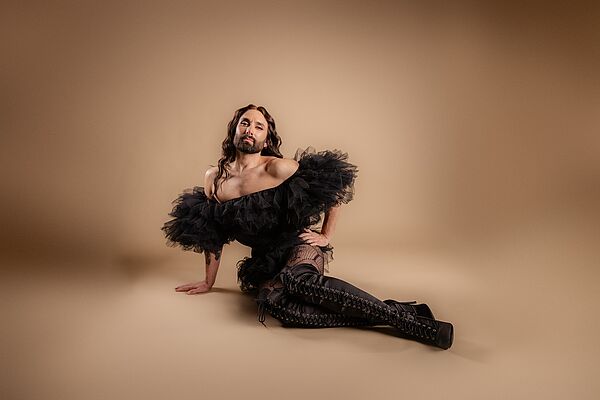 Conchita Wurst in einer schwarzen Abendrobe. Conchita Wurst sitzt auf der Erde.
