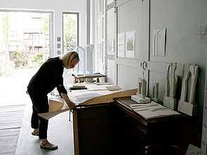 Julia Hansen breitet ein großes Stück Papier auf einem Tisch aus. Im Hintergrund stehen Skulpturen.