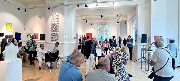 Menschen stehen und sitzen in einem Ausstellungsraum mit weißen Wänden, an denen Bilder hängen.