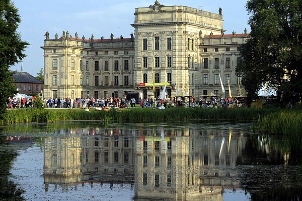 Ein Teich im Schlossgarten. Im Hintergrund steht das Ludwigsluster Schloss. Davor befinden sich viele Menschen. 