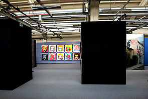 Blick in den Ausstellungsraum. Im Hintergrund hängen bunte Bilder.