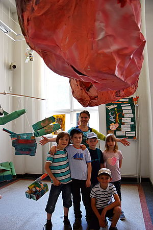 Die Künstlerin steht mit fünf Kindern unter einem roten Kunstobjekt, das wie ein Meteorit von der Decke hängt.