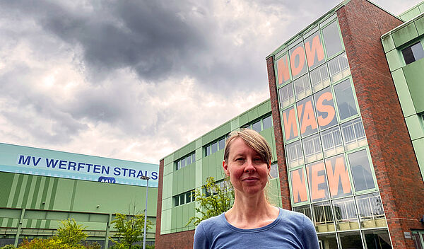 In den Scheiben der Glasfassade steht in orangener Farbe „Now was new“. Die drei Wörter stehen untereinander. Jedes Fensterelement enthält einen Buchstaben, der ungefähr so groß ist wie ein Erwachsener. Am Gebäude daneben prankt der Schriftzug „MV Werften Stralsund“. Im Vordergrund des Gebäudeensembles steht die Künstlerin. 