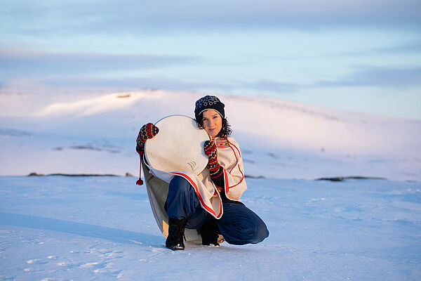 Eine Frau. Die Frau hockt im Schnee. Sie trägt eine Mütze, Handschuhe und einen Poncho. In der Hand hält sie ein rundes Musikinstrument. Das ist eine Rahmentrommel.