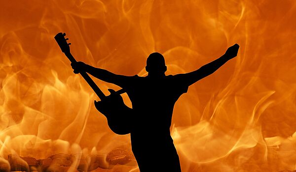 Vor orangem Hintergrund steht die Shilouette eines Mannes, der die Arme nach oben reißt und eine Gitarre in der Hand hält. 