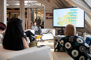 Martin Auer sitzt auf einem Sofa. Menschen hören ihm zu. 