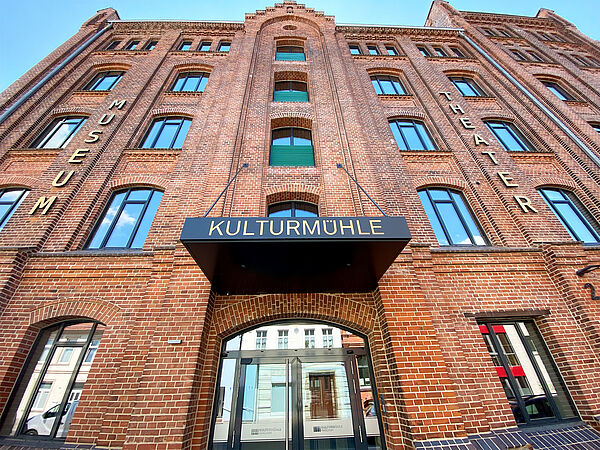 Die Backsteinfassade reicht fünf Stockwerke hoch. Etwa 20 hohe Fenster prägen das Bild. Links steht in großen goldenen Buchstaben Museum an der Fassade, rechts Theater. In der Mitte, über dem Glaseingang, hängt die Aufschrift Kulturmühle. 