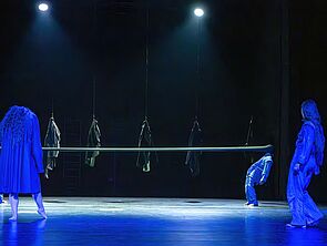 Eine blau gefärbte Bühne. An Haken hängen Puppen.