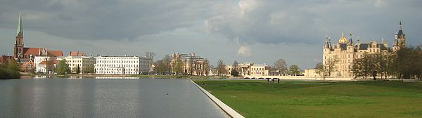 Das Panorama startet links mit dem Schweriner Dom, zieht an der weißen Staatskanzlei und dem beigefarbenen Theater vorbei bis zum Schloss. Im Vordergrund: das Wasser aus dem Burgsee und eine Wiese aus dem Schlossgarten. 