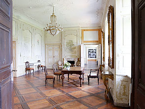 Ein Raum im Schloss. An der Decke hängt ein Kronleuchter. Im Raum stehen alte Stühle, zwei Tische und ein Kamin. An den Wänden scheinen alte Wandgemälde durch.