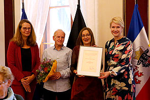 Ein Mann und eine Frau stehen zwischen Kulturministerin Bettina Martin und Ministerpräsidentin Manuela Schwesig und halten eine Urkunde sowie Blumen in den Händen.