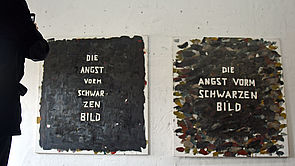 An der Wand hängen zwei dunkle Gemälde. Auf beiden steht - anders angeordnet - in weißer Schrift: Die Angst vorm schwarzen Bild.