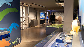 Links eine Wand mit bunten Sauriern. Im Hintergrund eine Ausstellungshalle.