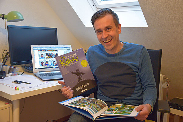 Stefan Pede sitzt vor seinem Schreibtisch, schaut in die Kamera und hält zwei seiner Bücher in der Hand. 