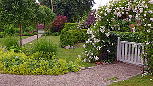 Ein Garten mit Wiesen, Bäumen, Blumen, Gartenwegen und einem weißen Gartentor.