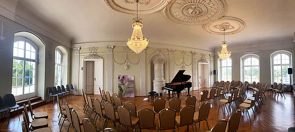 Ein Saal. Zwei Kronleuchter hängen von den Decken. Im Raum stehen Stühle und ein Klavier. Durch fünf hohe Fenster fällt Licht.