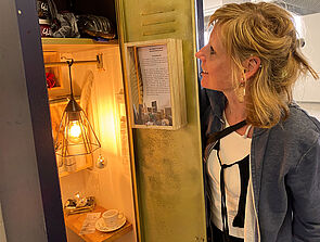 Ein geöffneter Spind. An der Decke brennt eine Lampe. Auf einem schmalen Brett stehen eine Tasse und eine Deko-Schale. An der Rückwand hängt ein Regal. Darauf stehen Gefäße und Stifte. Die Künstlerin blickt in den Spind.