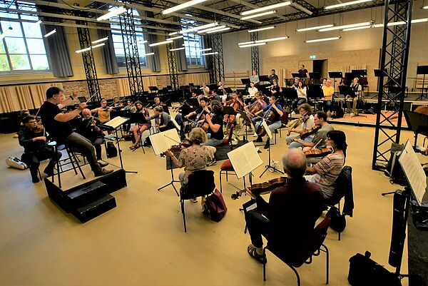 Alle Musikerinnen und Musiker haben an ihren Instrumenten Platz genommen. Links, auf einem Podest, sitzt Generalmusikdirektor Mark Rohde mit einem Taktstock in der Hand auf einem Hocker und gibt den Ton an. Das Orchester sitzt in einem Halbkreis vor ihm und spielt.