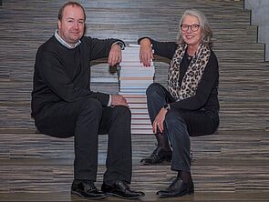 Ein Mann und eine Frau sitzen auf einer Treppe. Zwischen ihnen steht ein Stapel Bücher.