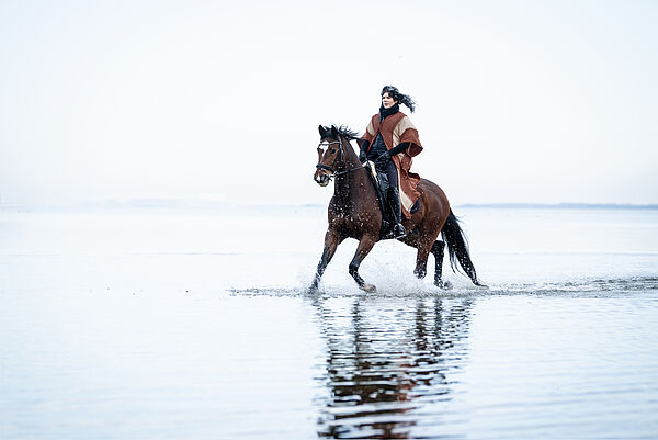 Eine Frau reitet auf einem Pferd durch flaches Wasser.