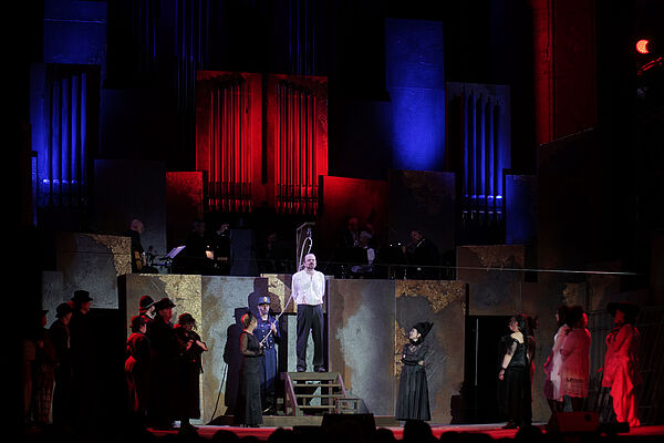 Eine Bühne. Auf der Bühne stehen viele Menschen. Das Licht ist auf einen Mann gerichtet. Der Mann steht unter einem Galgen. Die Schlinge liegt schon an seinem Hals.