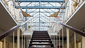 Ein Treppenhaus. Eine Treppe führt nach oben. Auf eine Ebene mit vielen Zellentüren.