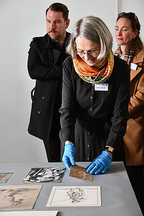 Eine Frau mit grauem Haar, orangefarbenem Schal und blauen Schutzhandschuhen beugt sich über einen Tisch und bewegt grafische Arbeiten. Im Hintergrund beobachten zwei weitere Personen die Szene. Auf dem Tisch liegen mehrere Druckgrafiken und Zeichnungen.