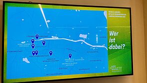 An einer Wand hängt ein Flachbildschirm mit einer Landkarte. Einige Orte sind mit Pins markiert.