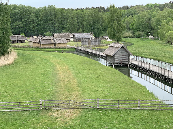 Auf einer Wiese stehen Hütten. Der Weg zu ihnen führt über einen schmalen Steg, der im Wasser steht.