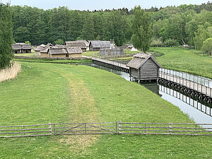 Auf einer Wiese stehen Hütten. Der Weg zu ihnen führt über einen schmalen Steg, der im Wasser steht.