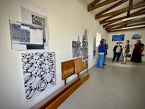 Der Ausstellungsraum. Vorn hängen Bilder an der Wand. Hinten stehen ein paar Menschen.