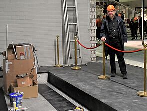 Generalintendant Hans-Georg Wegner trägt einen Bauhelm. Er steht vor einem roten Absperrband. Dahinter stehen Kartons und Farbeimer.