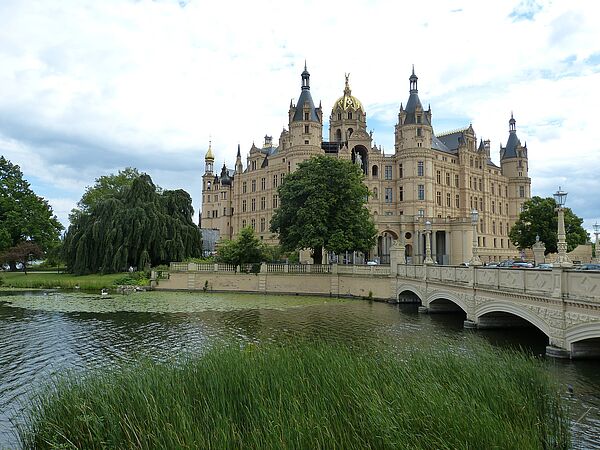 Das Schweriner Schloss mit Schlossbrücke. Im Vordergrund: hohe Gräser und der Burgsee.