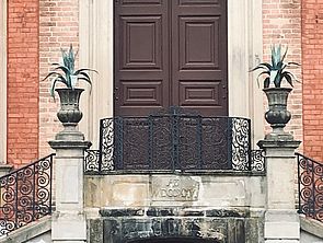 Die braune Eingangstür. Davor stehen zwei verzierte Säulen. Sie gehören zur Treppe, die zur Eingangstür führt.