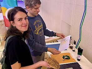 Eine Frau und ein Mann stehen an einem Tisch und setzen kleine Steine zu Bildern zusammen.