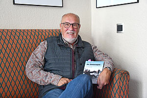 Ulrich Hinse sitzt auf einem Sofa und hält sein Buch in der Hand.