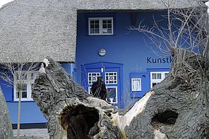 Hinter einem alten Baumstamm lugt der Ausschnitt eines blauen, restgedeckten Hauses hervor. Auf der Fassade steht "Kunstkaten".