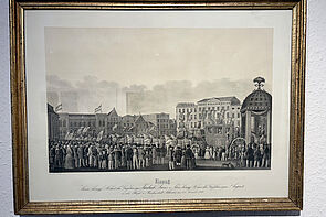 Ein Platz. Auf dem Platz stehen viele Menschen. Auf dem Platz steht eine Pferdekutsche. Im Hintergrund stehen Häuser.