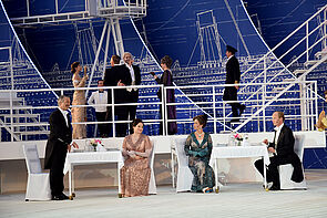 Ein Dinner an Deck. An zwei Tischen sitzen Paare. Rundherum stehen Gäste. 