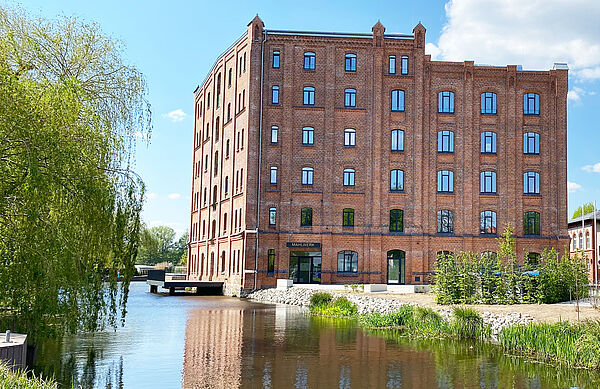 Die historische Fassade der ehemaligen Kornmühle zeigt sich von ihrer neuen Seite. Rund 80 Fenster prägend die beiden sichtbaren Seiten. Links vorm Gebäude fließt die Elde entlang. Im Fluss steht eine Terrasse. Sie gehört zum Restaurant der Kulturmühle. 
