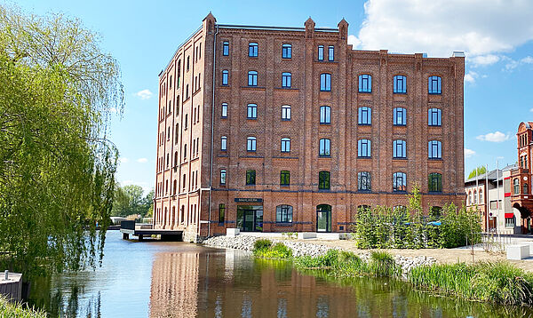 Die historische Fassade der ehemaligen Kornmühle zeigt sich von ihrer neuen Seite. Rund 80 Fenster prägend die beiden sichtbaren Seiten. Links vorm Gebäude fließt die Elde entlang. Im Fluss steht eine Terrasse. Sie gehört zum Restaurant der Kulturmühle. 