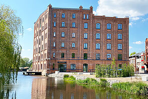 Die historische Fassade der ehemaligen Kornmühle zeigt sich von ihrer neuen Seite. Rund 80 Fenster prägend die beiden sichtbaren Seiten. Links vorm Gebäude fließt die Elde entlang. Im Fluss steht eine Terrasse. Sie gehört zum Restaurant der Kulturmühle. 